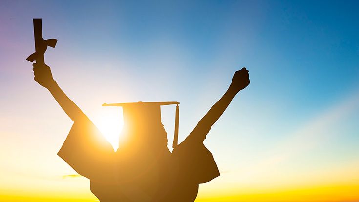 Silhouette of a person wearing graduation regalia raising their arms in celebration against a sunset background